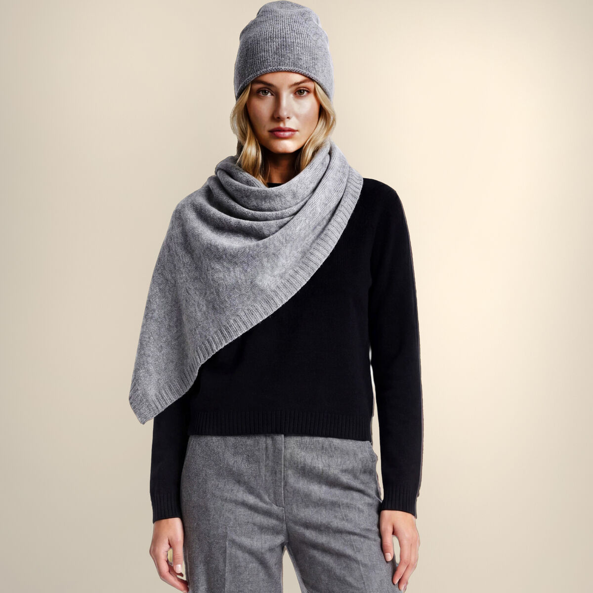 Eine Frau trägt eine naturbraune Kaschmirstola "Donna" locker um die Schultern, dazu eine farblich abgestimmte Mütze, schwarzen Pullover und weite graue Stoffhose. Der Hintergrund ist neutral beige, das Outfit wirkt elegant und gemütlich zugleich.