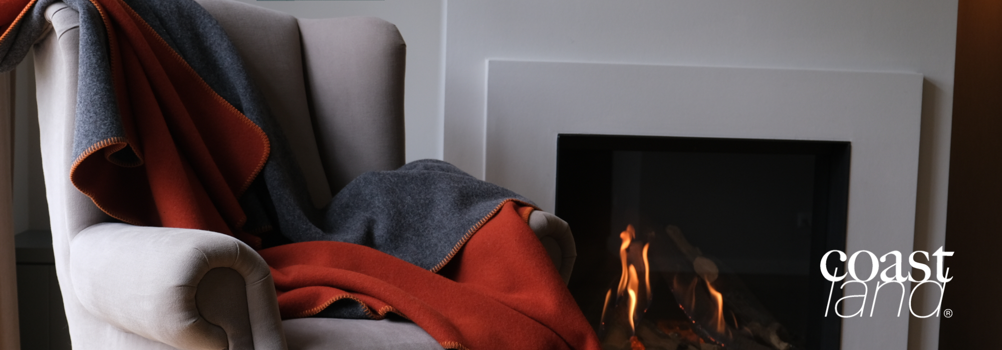Beige Sessel mit einer rot-grauen Decke, vor einem modernen Kamin mit flammendem Feuer. Logo von Coastland in der unteren rechten Ecke.