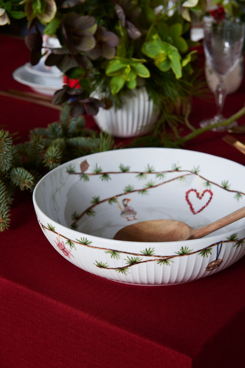 Weiße Schale mit weihnachtlichen Motiven, auf einem rot gedeckten Tisch, umgeben von grünen Pflanzen und Tannenzweigen.