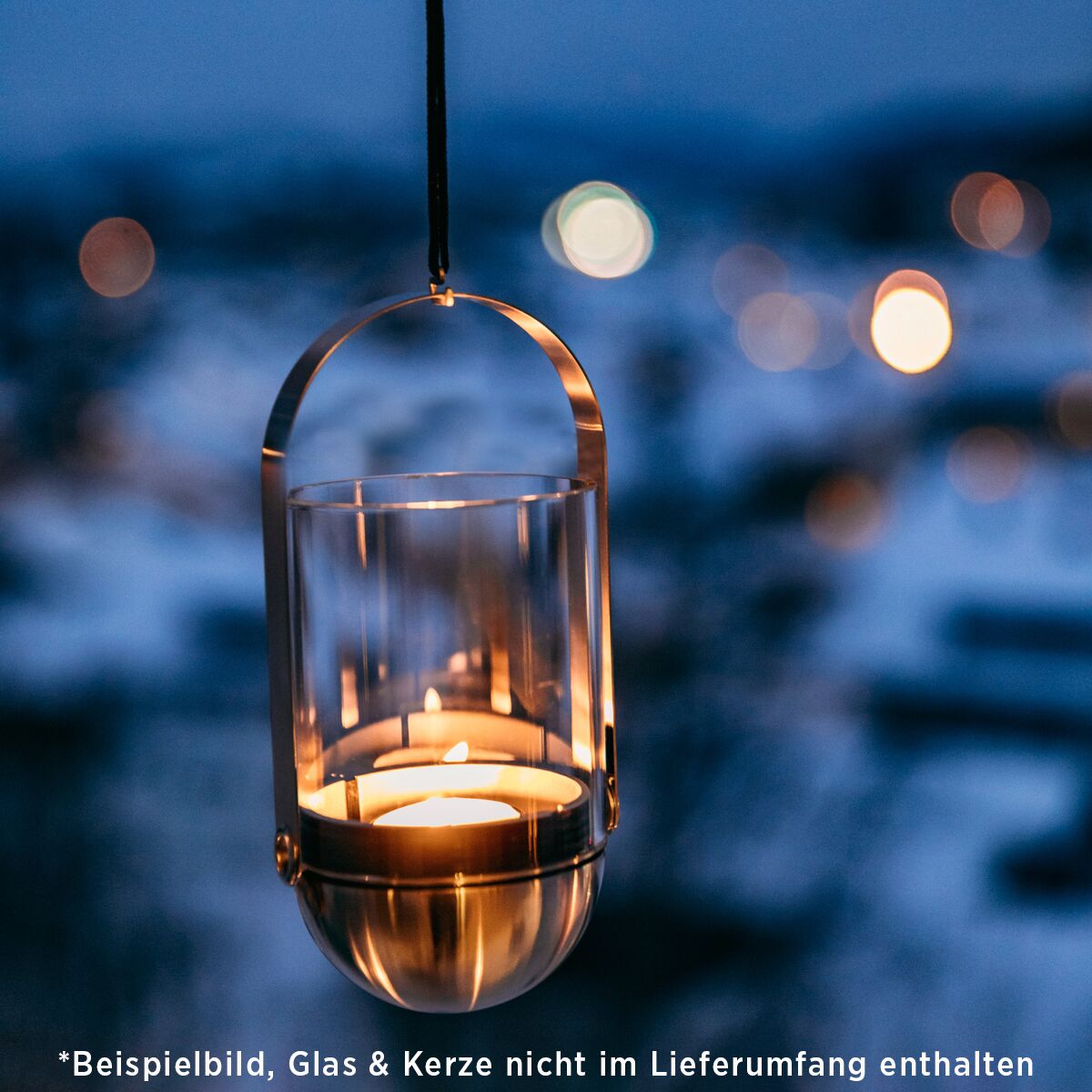 Ein modernes Hängesystem für Kerzen mit Glas, das draußen bei Dämmerung leuchtet. Die Metallhalterung hängt an einem schwarzen Seil, das Licht der Kerze verbreitet eine gemütliche und warme Atmosphäre. Im Hintergrund sind unscharfe Lichter und ein abendlicher Außenbereich zu erkennen.