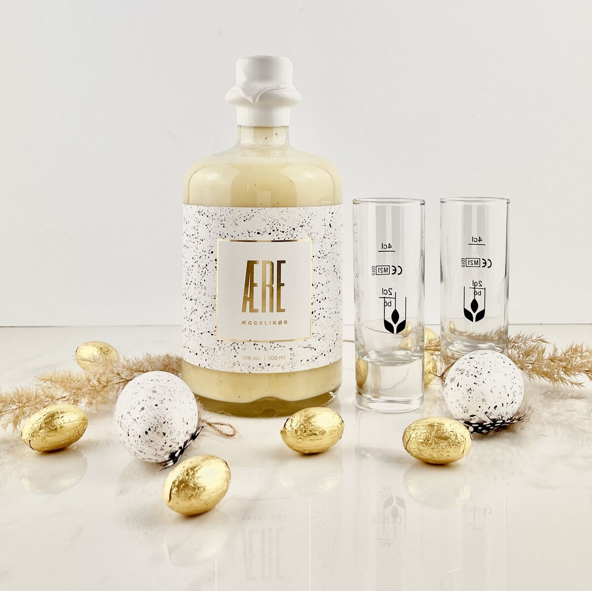 Eine Flasche Aere Eierlikör steht vor einem neutralen Hintergrund, flankiert von zwei durchsichtigen Gläsern und dekoriert mit weißen und goldenen Ostereiern.