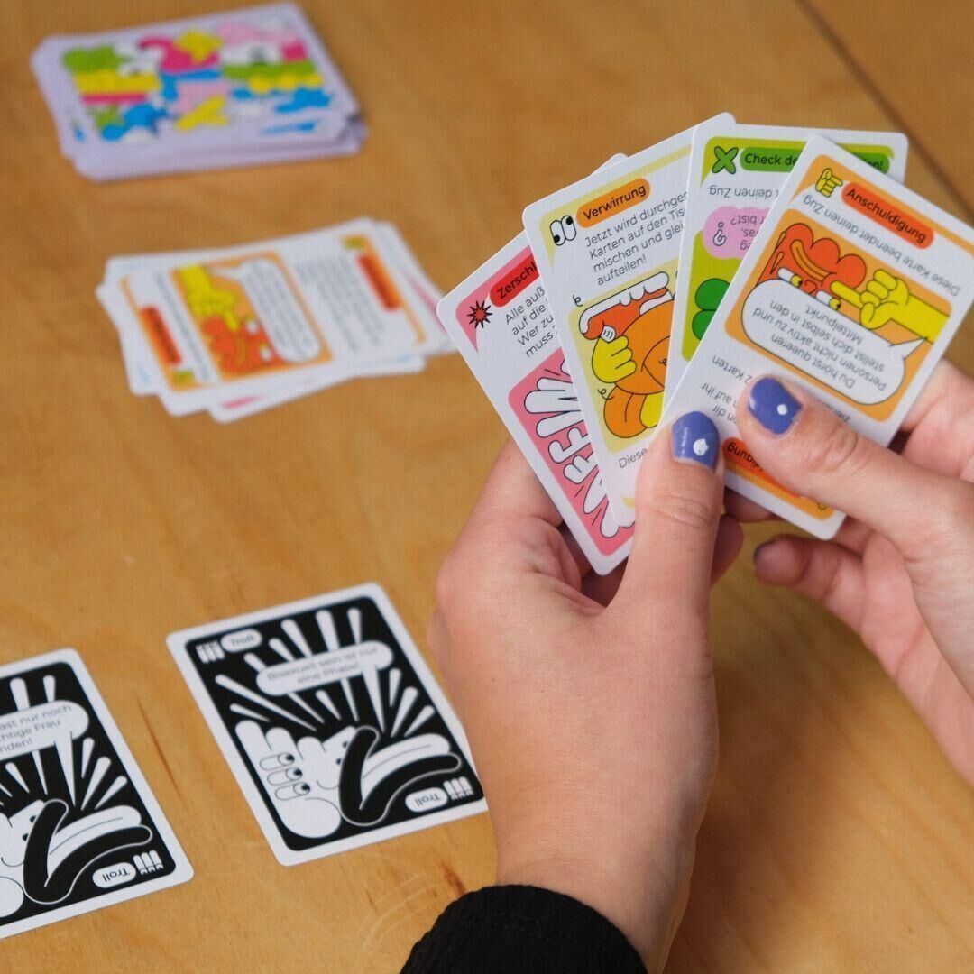 Personen spielen das Gesellschaftsspiel 'Queer Allyship - Das Kartenspiel'. Mehrere bunte Spielkarten, einige in der Hand einer Spielerin mit lila lackierten Nägeln, sind sichtbar. Auf dem Tisch liegen verschiedene Stapel von Spielkarten, darunter auch solche mit schwarz-weißen Designs.