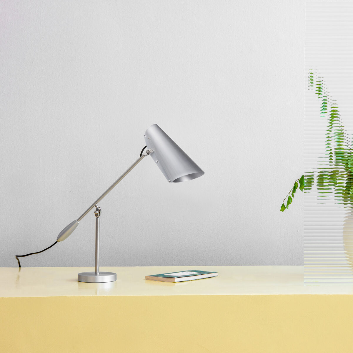 Eine moderne Tischlampe der Marke »Birdy« aus Metall in Silber. Die Lampe steht auf einem beigen Tisch neben einer grünen Pflanze in einem weißen Topf und einem Smartphone.