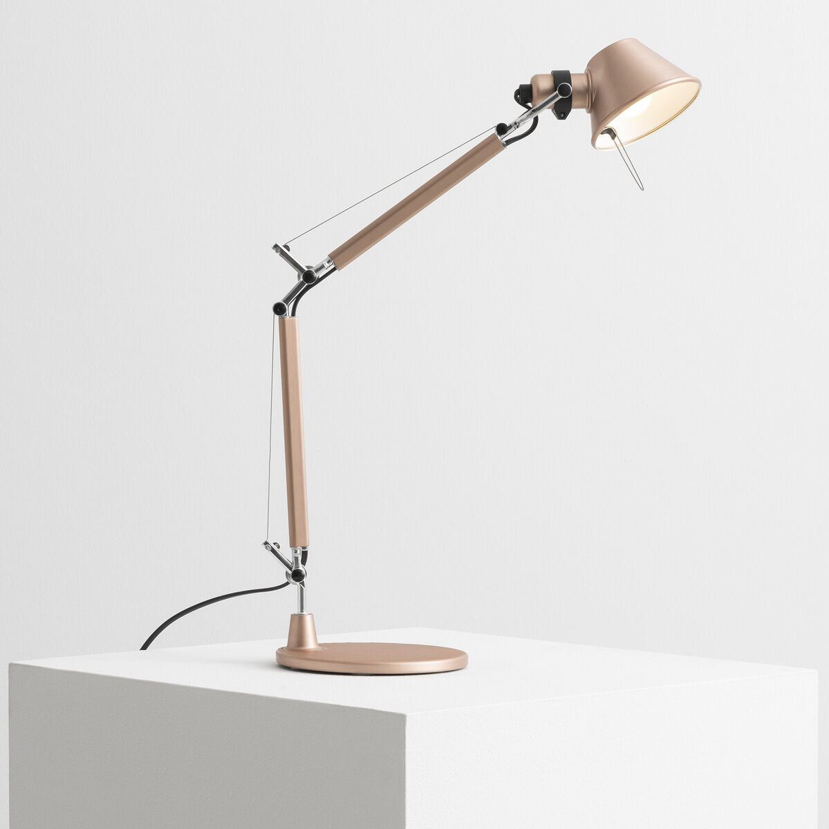 Die Artemide »Tolomeo Micro« ist eine schlanke, elegante Tischlampe in Beige und metallischen Farbtönen. Sie steht auf einem weißen Podest und hat einen verstellbaren Arm sowie einen schlichten runden Lampenschirm.