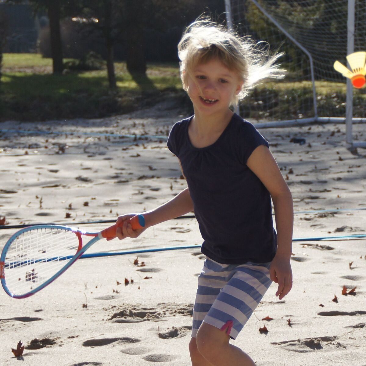 Ein Kind spielt am Strand mit einem Speedminton-Fun-Set. Es hält einen Schläger in der Hand und schaut lächelnd in die Kamera. Im Hintergrund sind ein Netz und Bäume zu sehen.