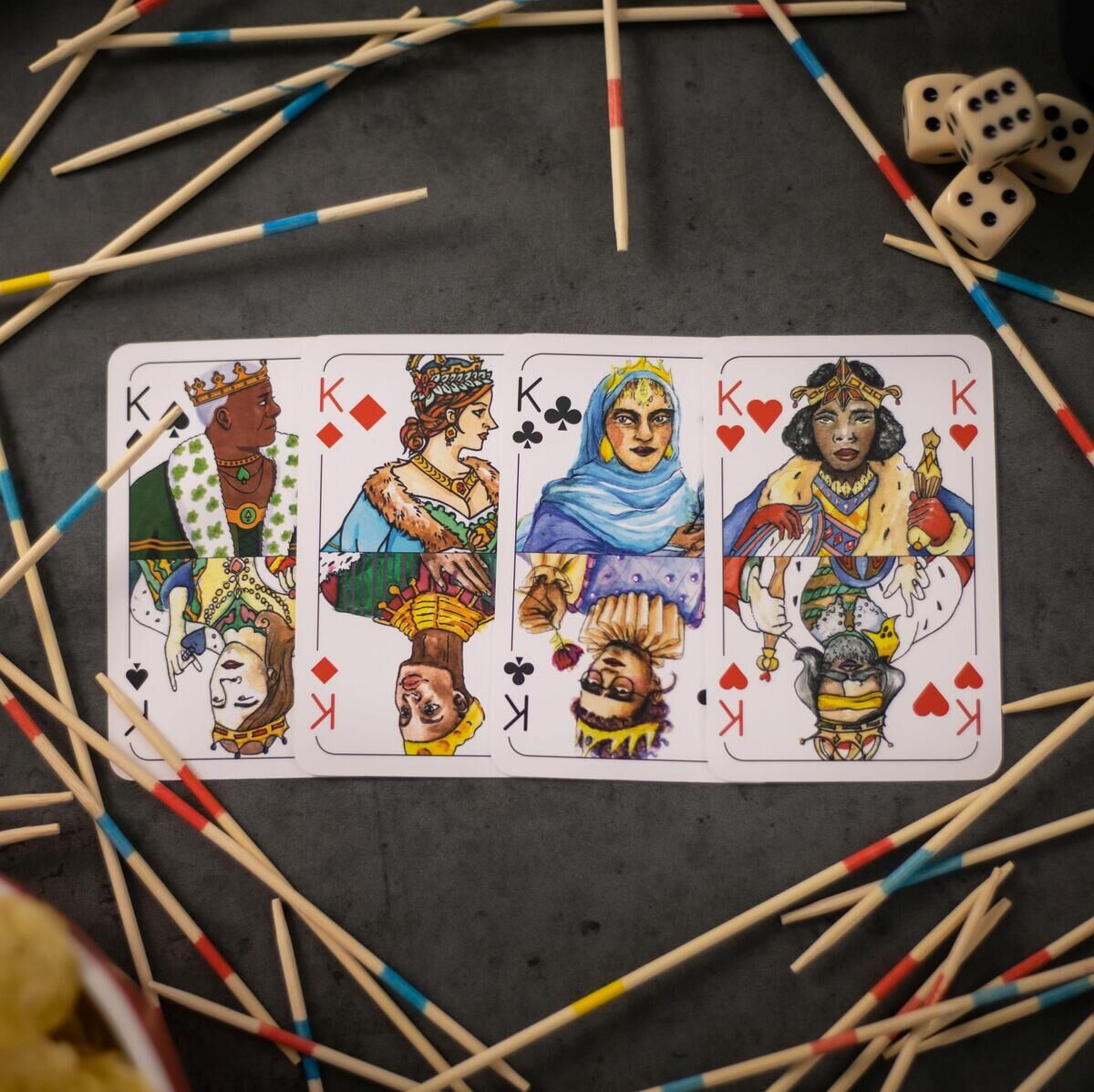Spielkarten für das Doppelkopf-Spiel liegen auf einem Tisch, umgeben von Mikadospieße und Würfeln. Vier Königskarten mit bunten Illustrationen sind zu sehen. Neben den Karten ist ein Schälchen mit Chips.