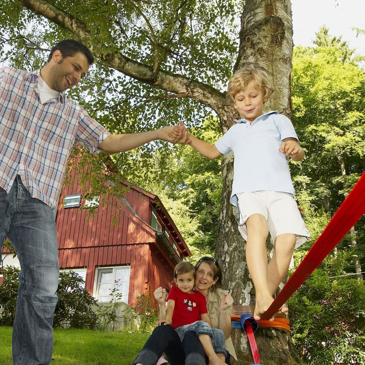 Ein Mann hält die Hand eines jungen Jungen, der auf einer blauen Slackline balanciert. Im Hintergrund sind zwei Frauen und ein weiteres Kind auf der Wiese zu sehen. Bäume und ein rotes Haus sind ebenfalls im Hintergrund sichtbar.