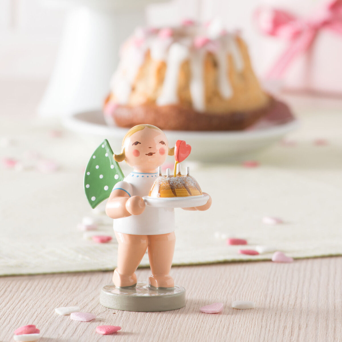 Im Vordergrund steht eine handbemalte Holzfigur eines Engels mit grün-gepunktetem Flügel, Kuchen und Herz von Wendt & Kühn. Dahinter auf weißer Tischdecke zwei Glasur-Gugelhupfe, eine rosa Geschenkbox und bunte Wimpelgirlande als Dekoration.