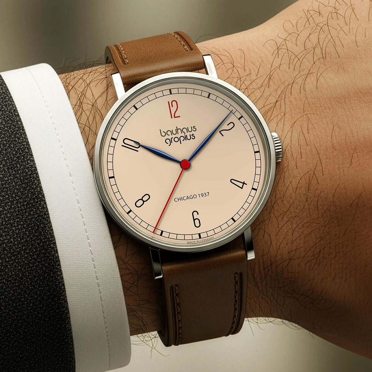 Foto einer stilvollen Armbanduhr der ZEIT-Sonderedition Walter Gropius »New Bauhaus Chicago 1937«. Die Uhr hat ein rundes silbernes Gehäuse, ein weißes Zifferblatt mit minimalistischer Gestaltung, filigrane blaue und rote Zeiger sowie ein braunes Lederarmband. Sie wird an einem Handgelenk vor einem neutralen Hintergrund getragen.