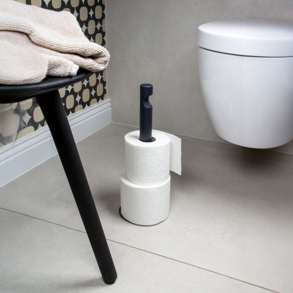 Modernes Bad mit mattschwarzem, freistehendem Rollenhalter: Auf dem runden Fuß stehen zwei weiße Toilettenpapierrollen, die obere ist auf den schlanken Stab gesteckt. Daneben ein wandhängendes weißes WC, graue Fliesen, ein schwarzer Hocker mit beigen Handtüchern und gemusterte Tapete.