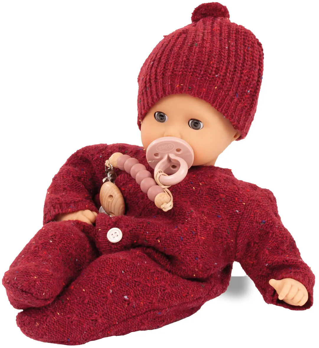 Babypuppe »Muffin Baby Jubilee« trägt einen bordeauxroten Strickanzug mit passender Mütze und hält einen rosa Schnuller mit Perlenkette in der Hand. Die Puppe sitzt leicht angelehnt und wirkt weich und kuschelig. Im Hintergrund ist ein neutraler weißer Untergrund zu sehen.