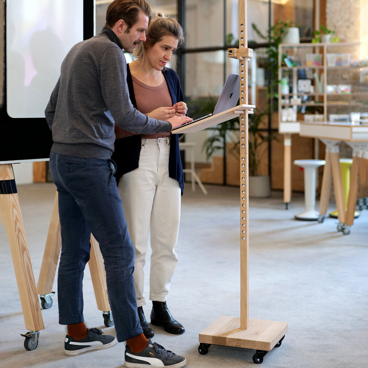 Zwei Personen arbeiten gemeinsam an einem Laptop, der auf dem mobilen Device Stativ »ERA« aus hellem Eichenholz steht. Das Stativ besitzt Rollen, ist höhenverstellbar und steht in einem modernen, hellen Raum mit weiteren Einrichtungsgegenständen und Pflanzen.