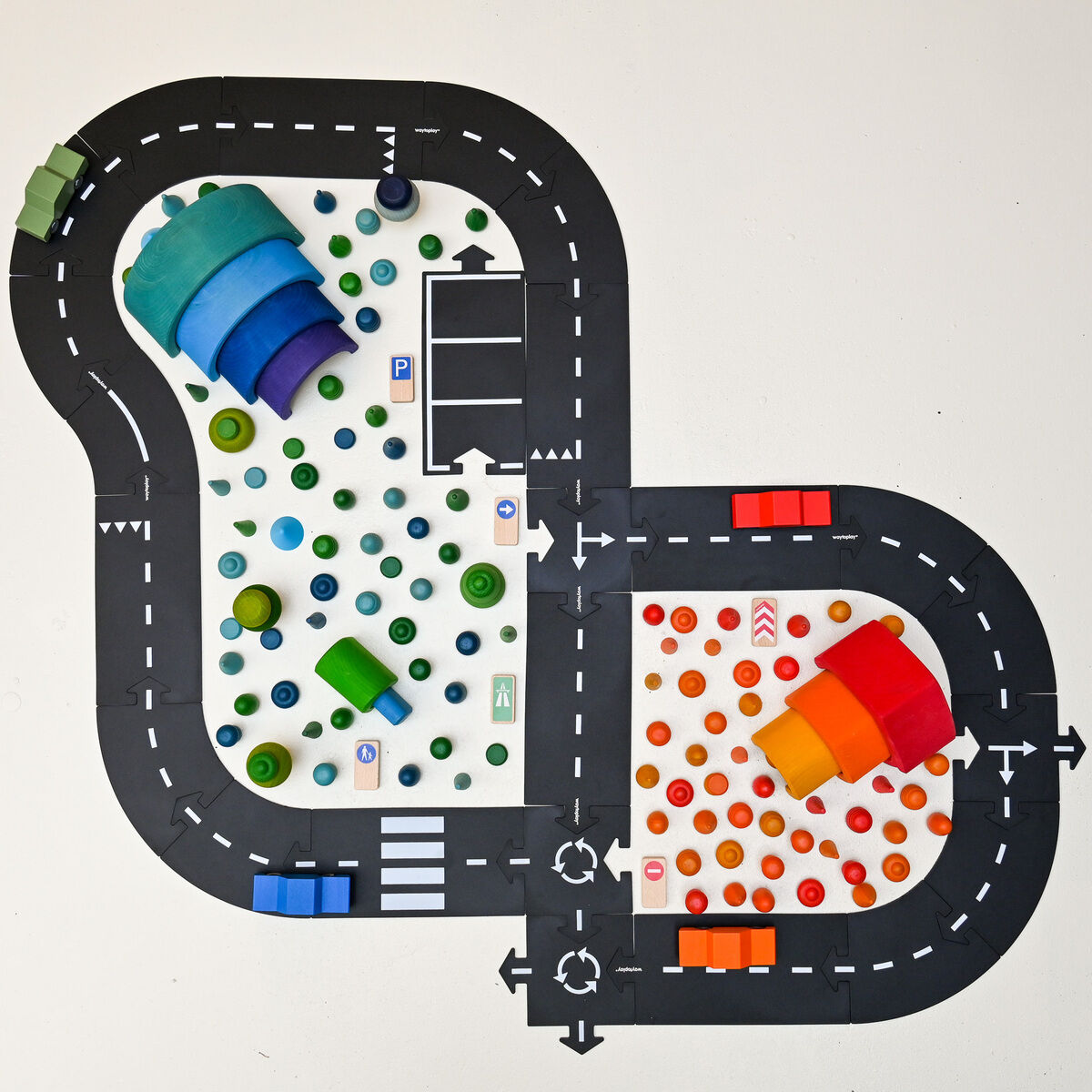 Spielset für Kinder mit schwarzer Straßenbahnmatte und bunten Holzbausteinen, angeordnet auf einer weißen Fläche. Das Set umfasst Fahrbahnen, Parkplätze und farbige Häuser, ideal zur Förderung von Kreativität und Verkehrserziehung.