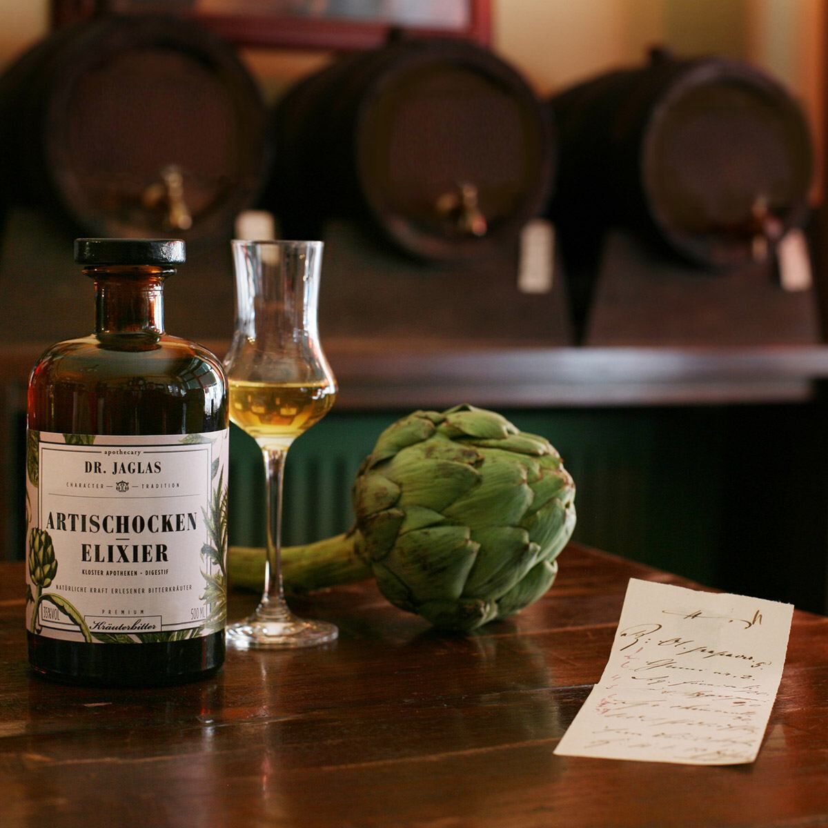 Eine Flasche »Artischocken-Elixier« von Dr. Jaglas steht auf einem Holztisch neben einem Glas gefüllt mit dem Kräuterbitter und einer ganzen Artischocke. Im Hintergrund sind mehrere Holzfässer zu sehen. Auf dem Tisch liegt ein handgeschriebener Zettel.