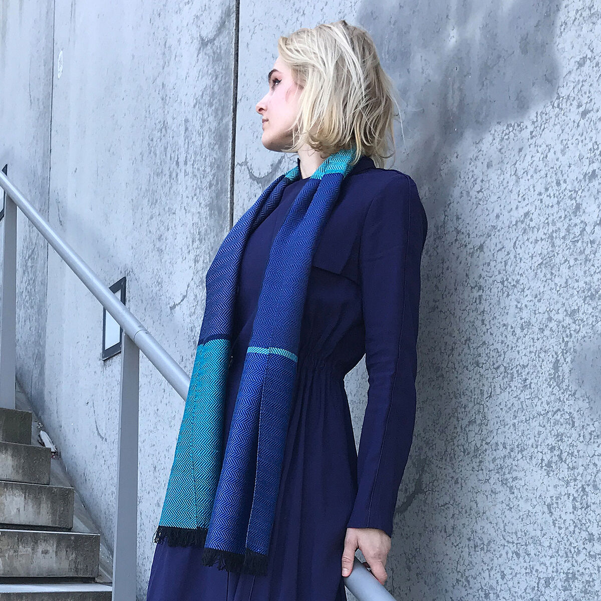 Blonde Frau steht auf einer Treppe und trägt einen blauen und türkisfarbenen Seidenschal sowie ein dunkelblaues Kleid.