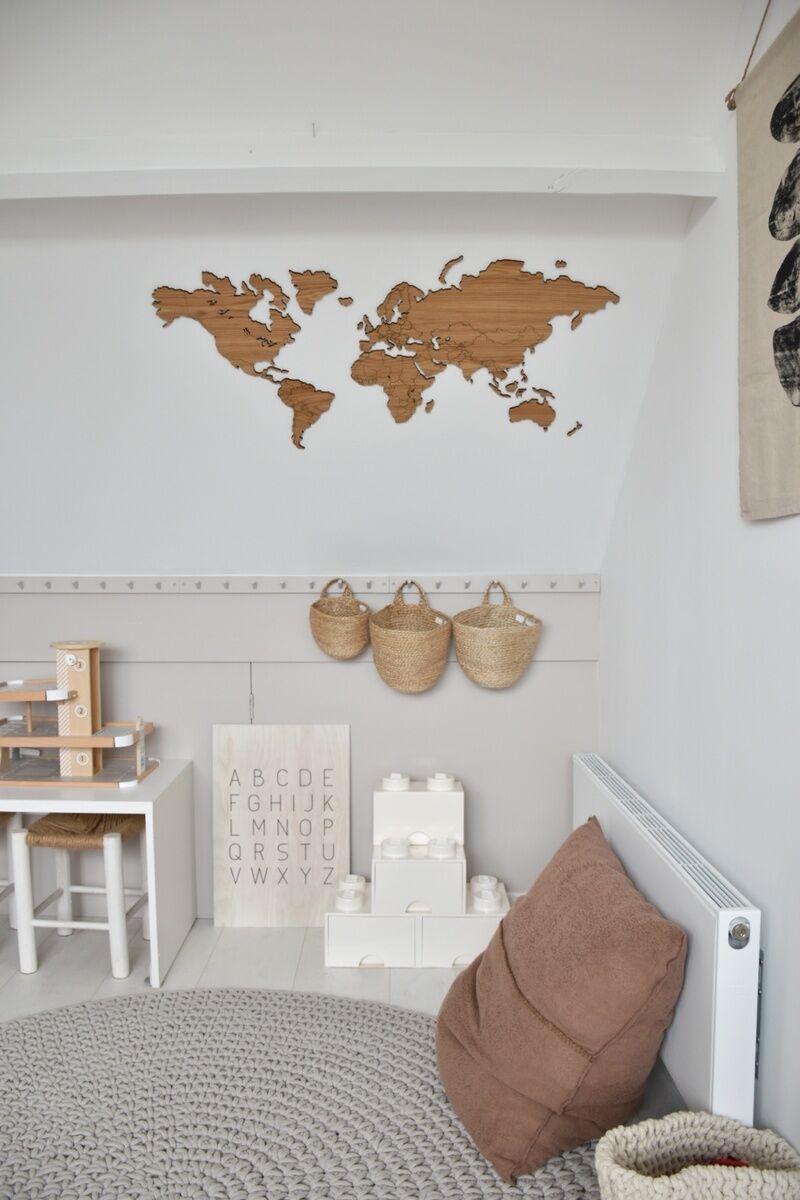 Selbstklebende Weltkarte aus Holz an einer weißen Wand angebracht, umgeben von minimalistischer Dekoration in einem modernen Raum mit einem Spieltisch, Korbhängern und einem großen Kissen.