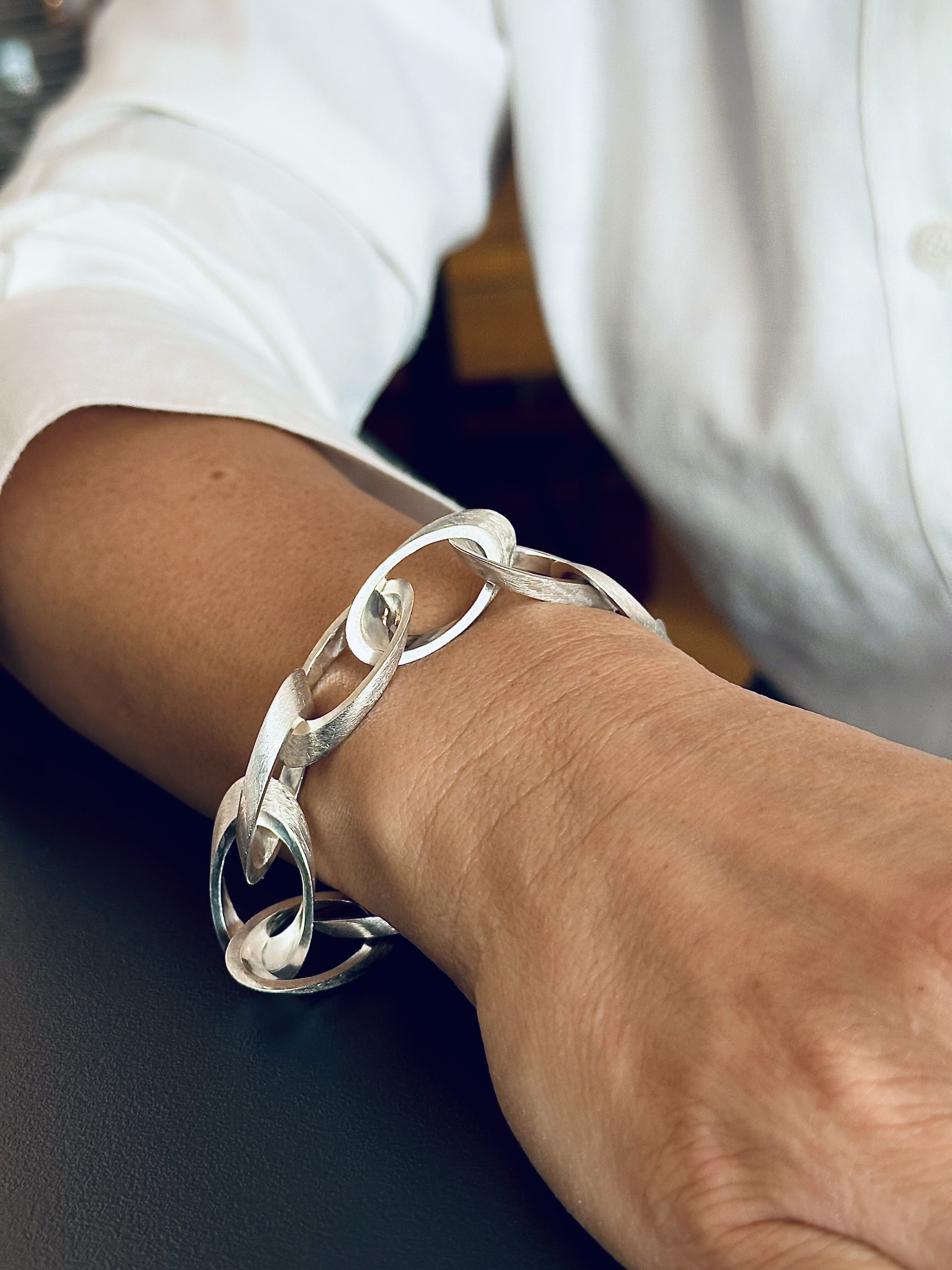 Ein stilvolles silbernes Armband mit einem elliptischen Design wird am Handgelenk einer Person getragen, die ein weißes Hemd trägt.