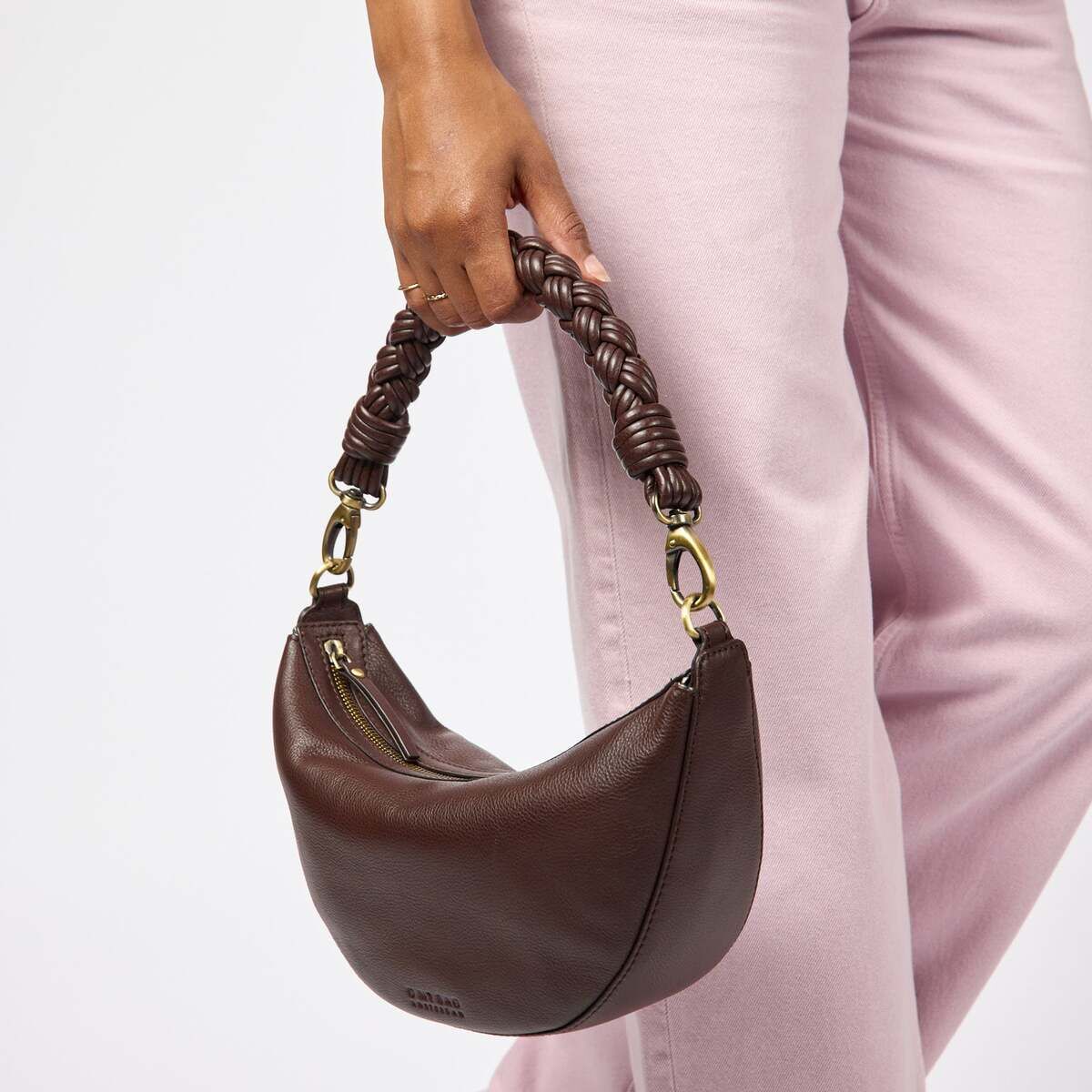 Eine Frau trägt eine mittelgroße, braune Handtasche »Leo« mit geflochtenem Henkel aus Lederoptik und goldfarbenen Details. Die Tasche wird in der Hand gehalten und passt zu einer hellrosafarbenen Hose und einem Pullover. Die Szene ist modisch und modern inszeniert.