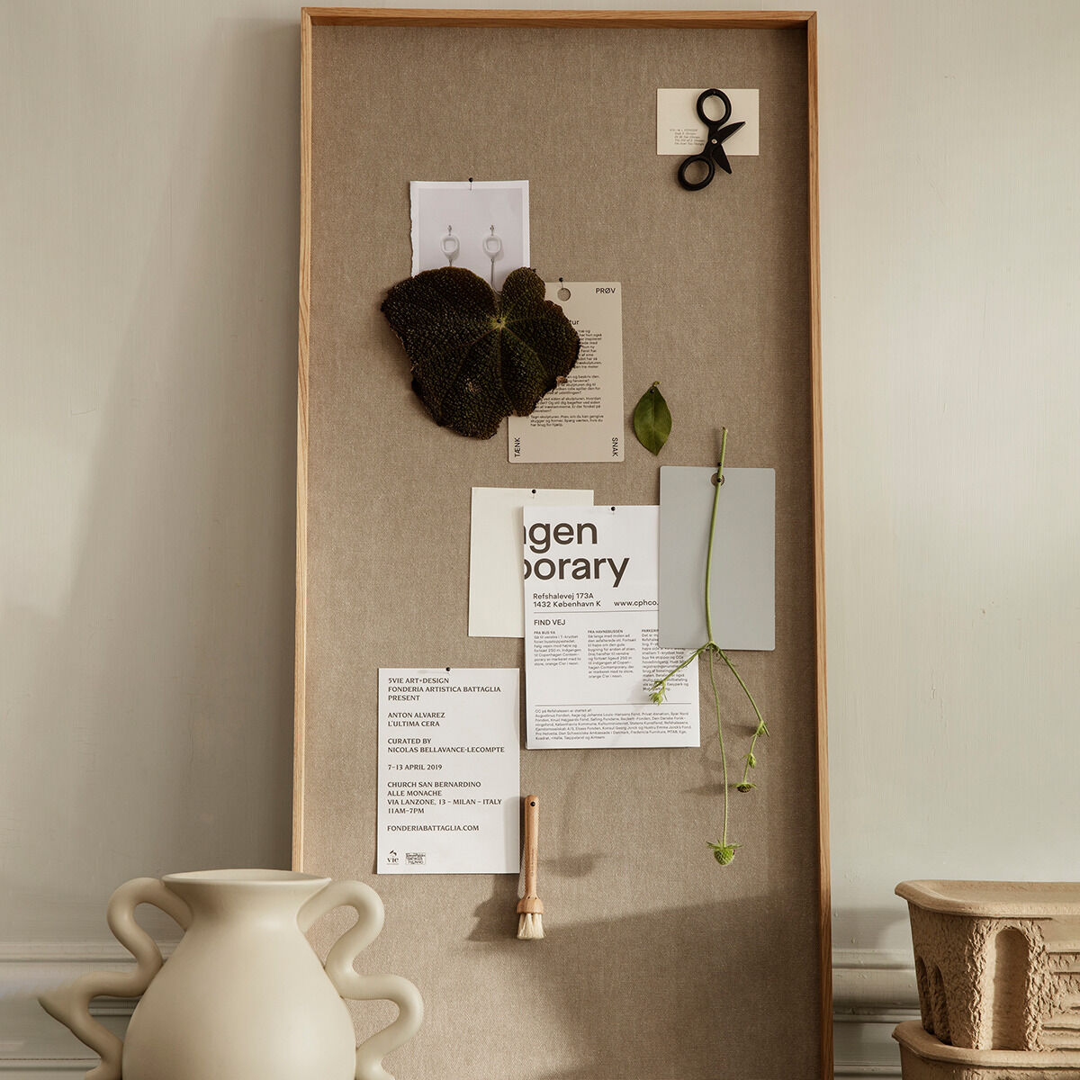 Eine minimalistische Pinnwand mit einem Holzrahmen ist an einer beigen Wand befestigt. Auf der Pinnwand sind verschiedene Notizen und ein kleiner grüner Zweig angebracht. Darunter steht eine weiße Vase.