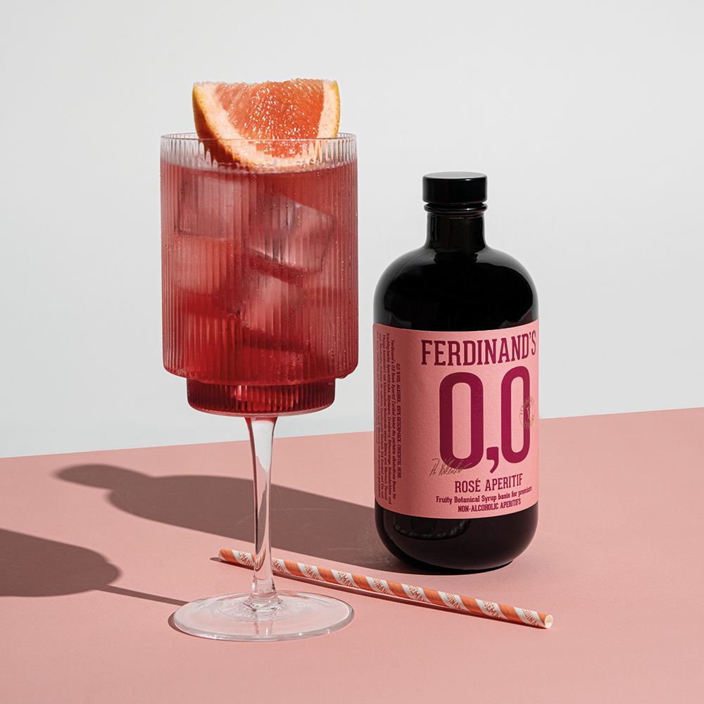 Glas mit rosafarbenem Aperitif, Eiswürfeln und Grapefruitscheibe neben Flasche Ferdinand's 0,0 Rose Aperitif und gestreiftem Trinkhalm