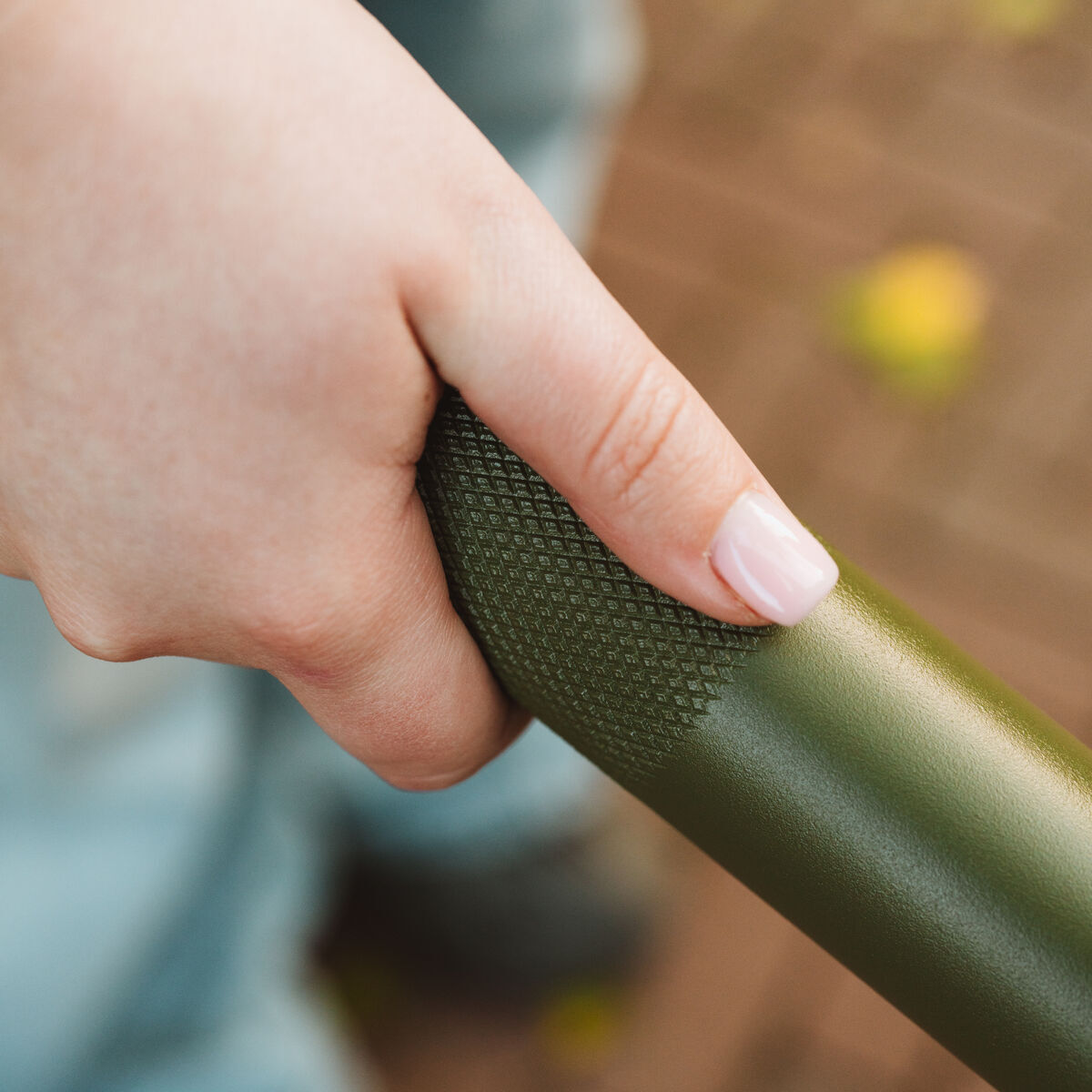 Detailaufnahme einer Hand, die einen grün lackierten Metallgriff mit rutschfester Struktur hält. Im Hintergrund sind verschwommen ein gepflasterter Boden, gelbe Blätter und blau gekleidete Beine zu sehen. Die Szene wirkt herbstlich und einladend.