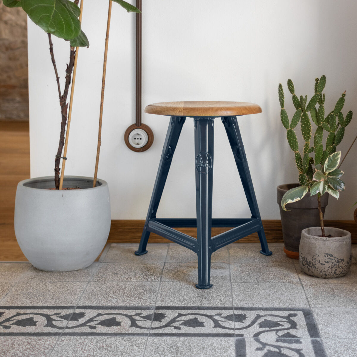 Ein stilvoller Rowac-Schemel in der Farbe 'Blue Moon' steht neben mehreren Zimmerpflanzen in Töpfen. Der Hocker hat einen hölzernen Sitz und blaue Metallbeine. Er ist vor einer weißen Wand und auf einem gemusterten Fliesenboden platziert.