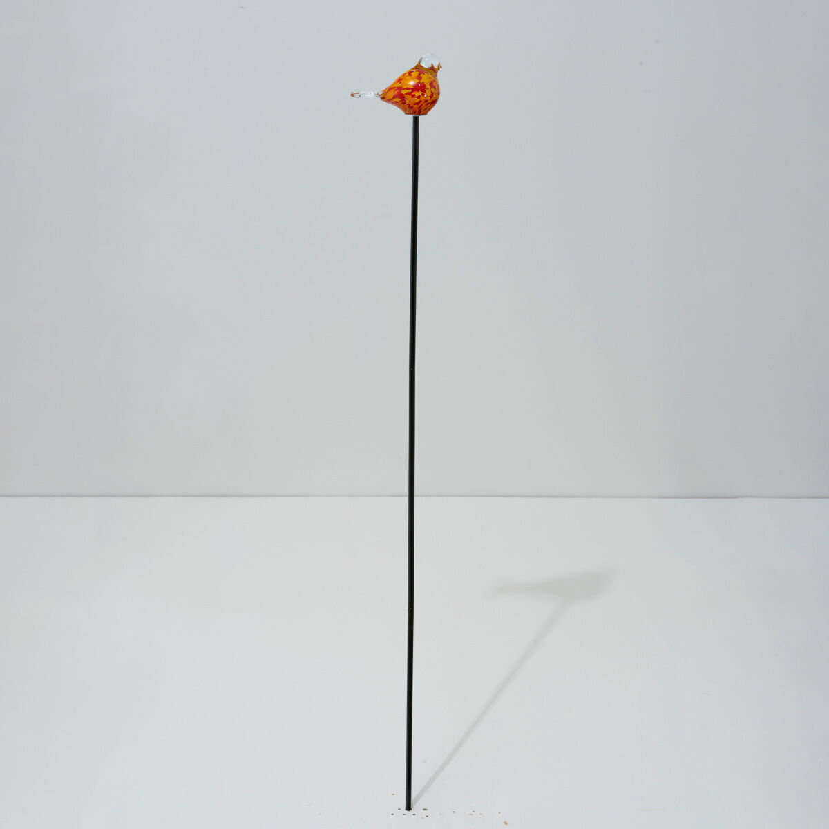 Moderner Gartenstecker namens "Gartenstecker Vogeltrio" der Marke Ars mundi, bestehend aus einem langen schwarzen Metallstab, an dessen Spitze ein orangefarbener, gläserner Vogel angebracht ist. Minimalistisches Design, auf hellem Hintergrund, geeignet für den Garten.