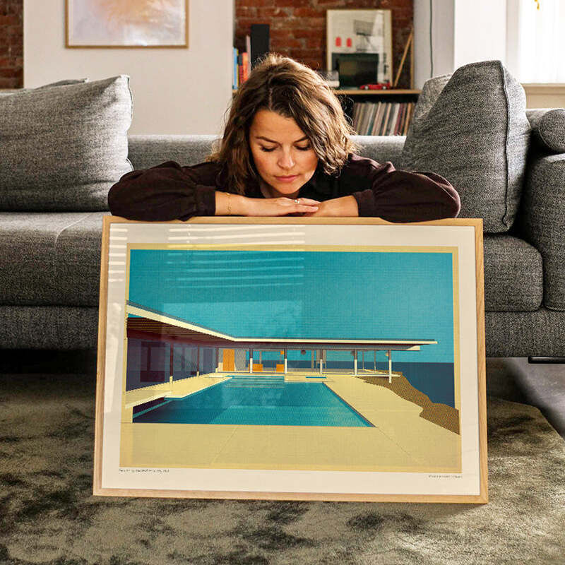 Frau liegt auf einem grauen Teppich vor einem Poster des Stahl Case Study House 1959 von Pierre Koenig mit Rahmen. Im Hintergrund befindet sich ein modernes Wohnzimmer mit grauem Sofa und Bücherregal. Das Poster zeigt ein modernes Haus mit Pool in klaren Blautönen und minimalistischer Architektur.