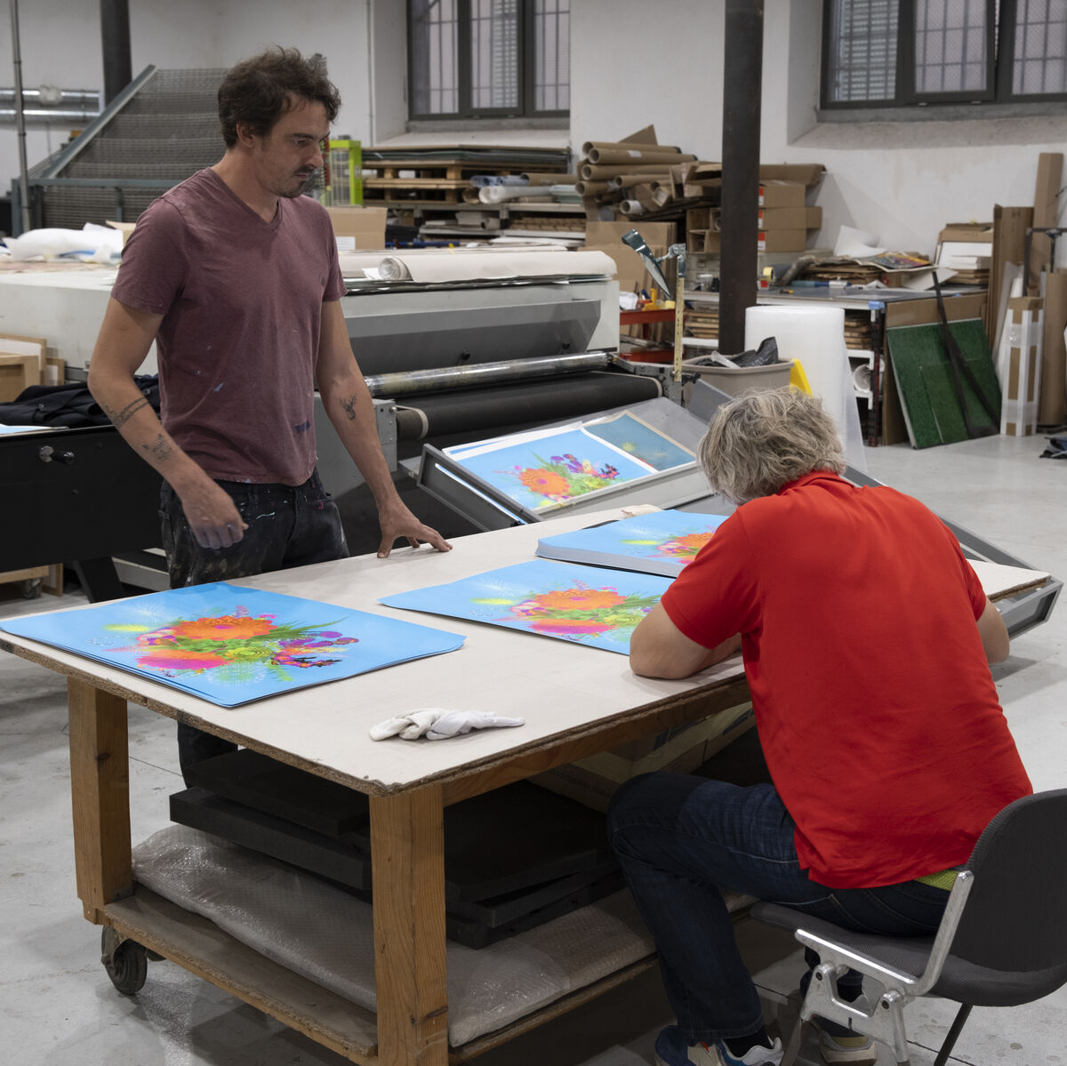 Zwei Männer arbeiten in einem Künstleratelier an mehreren farbenfrohen Kunstwerken. Der Raum ist mit Malutensilien und diversen Materialien ausgestattet.