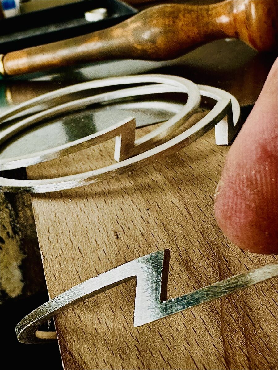 Detaillierte Nahaufnahme von mehreren silberfarbenen Armreifen mit markantem Z-förmigem Designelement auf einer Holzunterlage. Im Hintergrund Werkzeuggriff, Fokus auf die handgefertigte, exklusive ZEIT Sonderedition von HML Berlin.