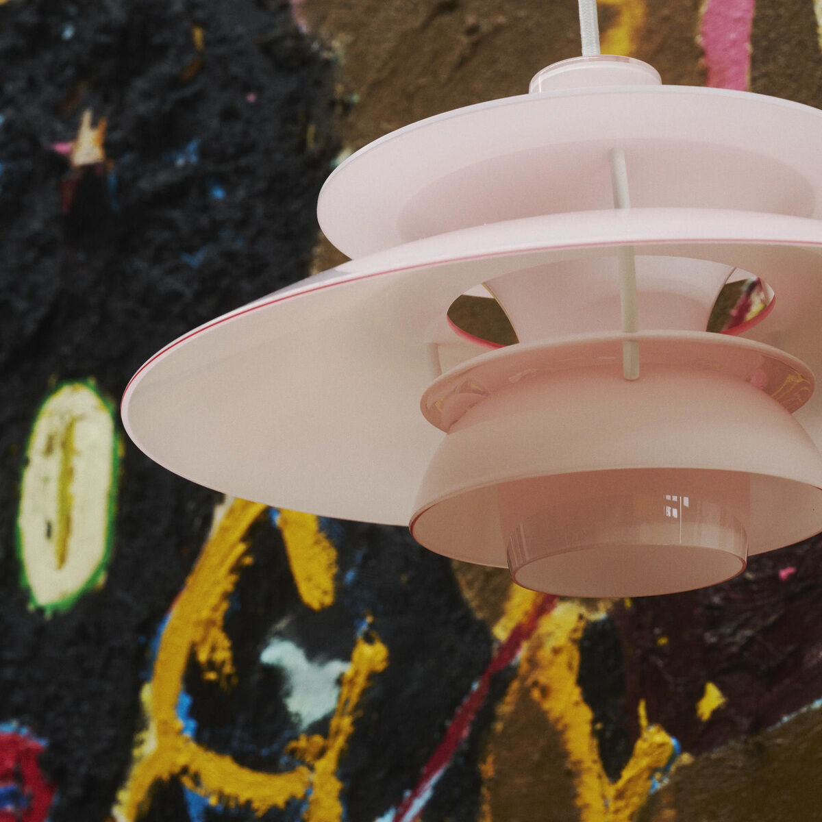 Detailaufnahme der Hängeleuchte »PH 5 320« von Louis Poulsen in Weiß und Pale Rose vor kunstvoll bemalter Wand. Die Leuchte aus glänzendem, pulverbeschichtetem Polymer zeigt ihr ikonisches, dreiteiliges Schirmsystem und sorgt für blendfreies Licht.