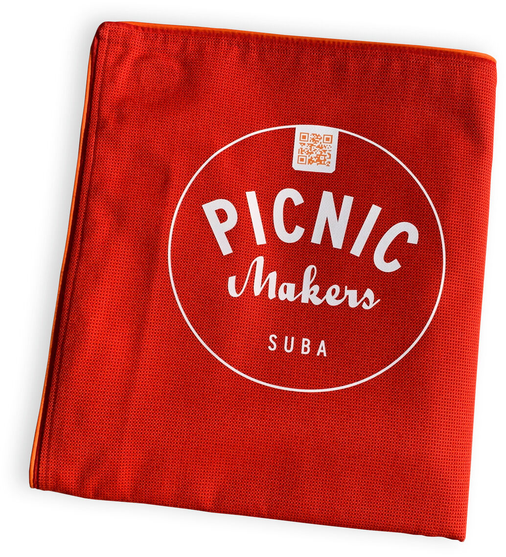 Quadratische, in Rot gehaltene Picknickdecke mit weißem Aufdruck. Die Decke ist kompakt zusammengefaltet und trägt die Aufschrift: 'PICNIC Makers SUBA'.