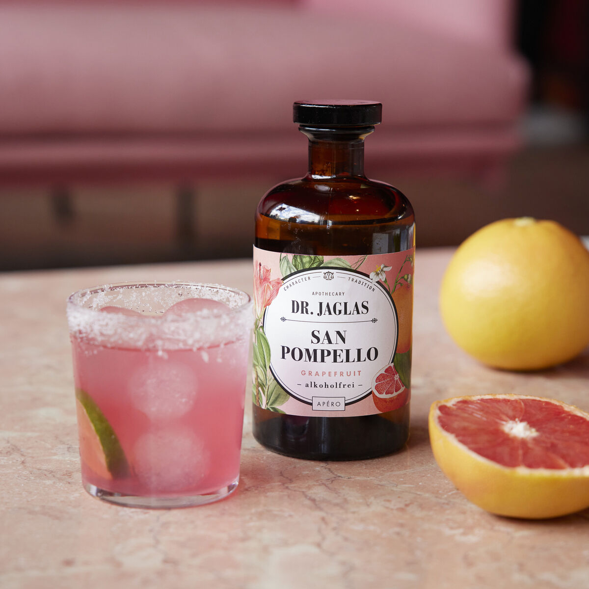 Flasche des alkoholfreien Aperitifs »San Pomello« von Dr. Jaglas auf einem Tisch. Daneben ein Glas mit pinker Flüssigkeit und Eiswürfeln sowie eine aufgeschnittene Grapefruit.