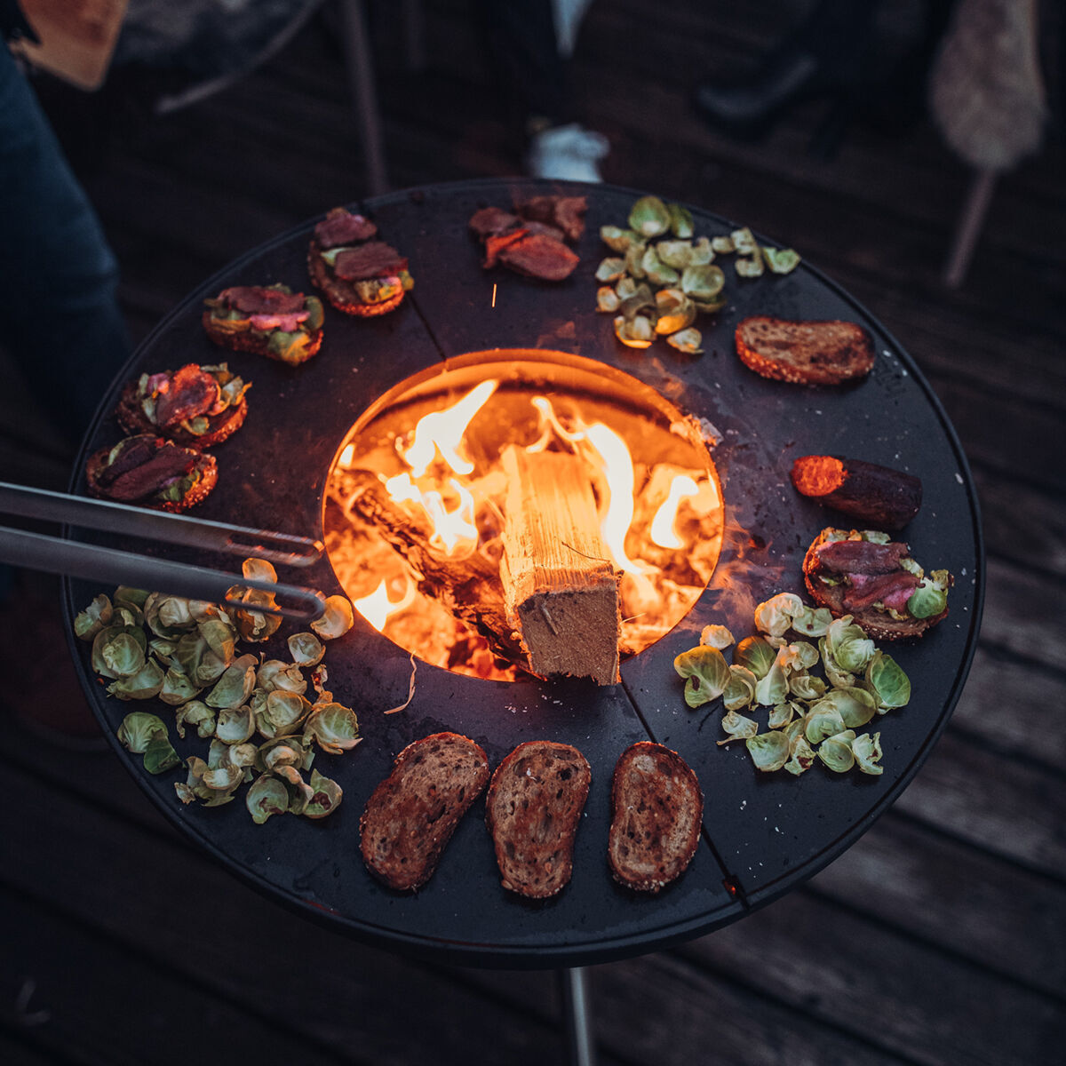 Eine »BOWL« Plancha Halbring ist im Einsatz. Auf dem kreisförmigen Metallkochfeld um ein Holzfeuer herum liegen verschiedene Grillspeisen wie Brot, Fleisch und Gemüse. Die Szene ist im Freien auf einer Holzterrasse.