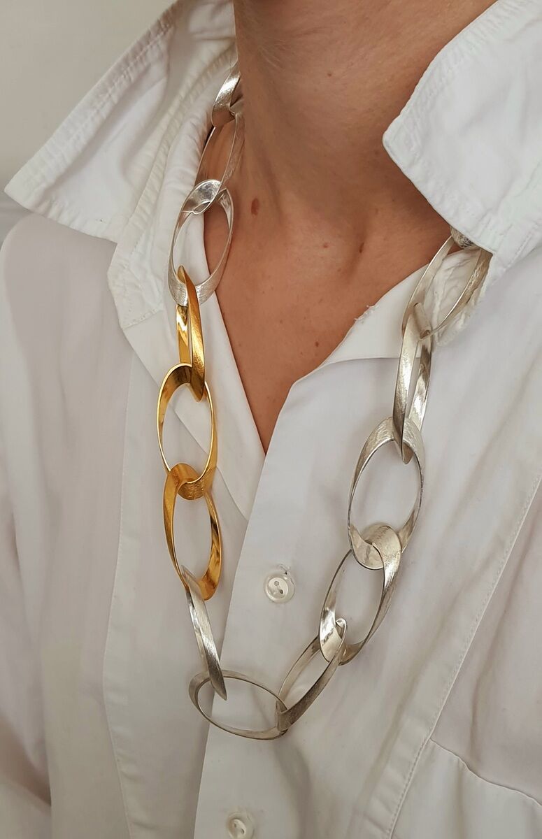 Ein elegantes Collier aus Silber und Gold, getragen von einer Person mit weißem Hemd. Die Kette besteht aus großen, ineinander verschlungenen Gliedern in metallischem Glanz, die sowohl goldene als auch silberne Elemente enthält.