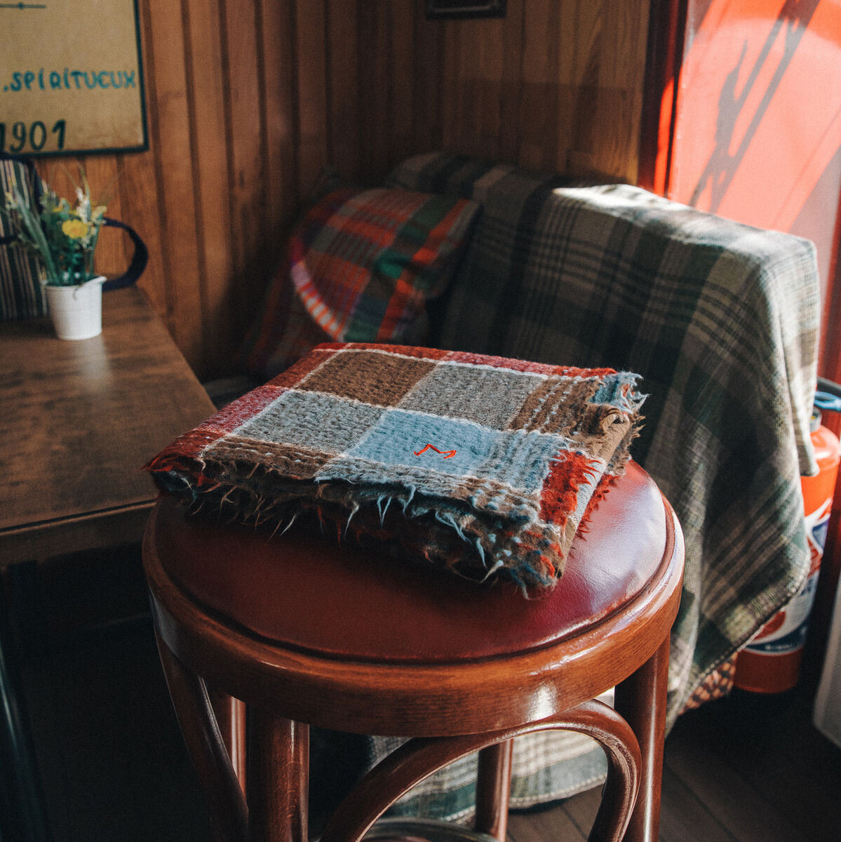 Ein Halstuch im Patchwork-Design liegt auf einem runden Holzhocker. Der Schal besteht aus verschiedenen Wollstücken in den Farben Braun, Rot, Blau, Grau und Orange. Im Hintergrund befindet sich ein Holzinterieur mit karierten Decken und Kissen.