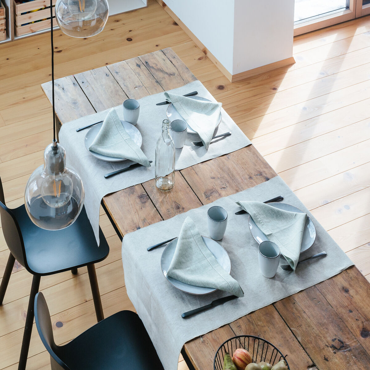 Ein natürlicher Leinen-Tischläufer auf einem rustikalen Holztisch, dekoriert mit weißem Geschirr und grauen Servietten, gesehen von oben in einem modern eingerichteten Esszimmer mit hellen Holzböden und einer Glasflasche in der Mitte.