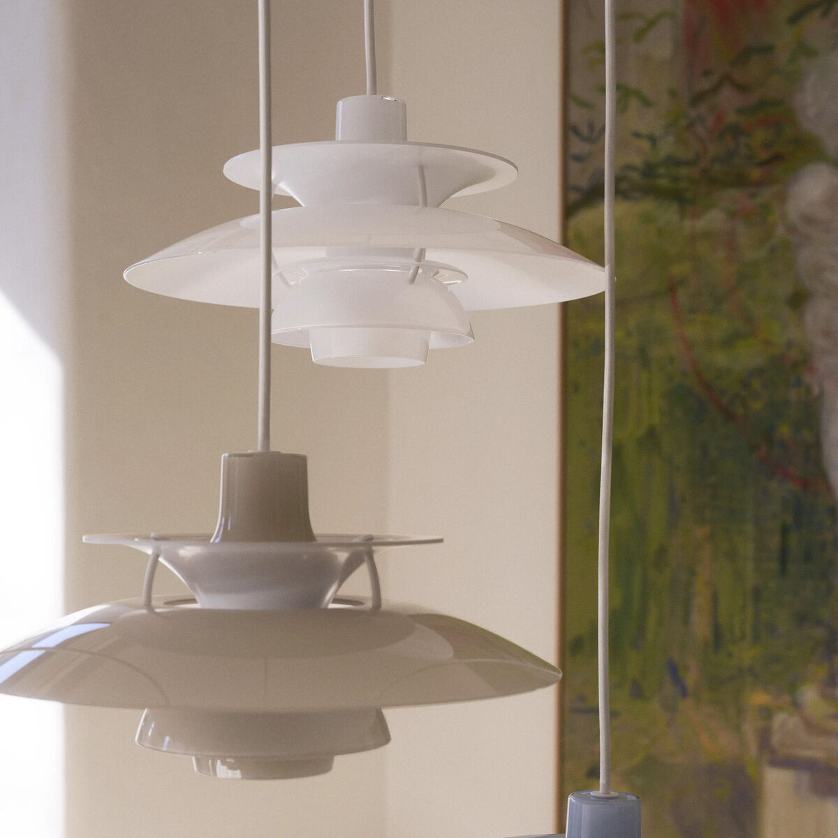 Zwei moderne, glänzend weiße Hängeleuchten im ikonischen Drei-Schirm-Design von Louis Poulsen hängen nebeneinander vor einer hellen Wand mit Kunstbild. Die opalen Leuchtenschirme streuen das Licht weich und blendfrei in den Raum. Zeitloses, elegantes Wohnaccessoire.