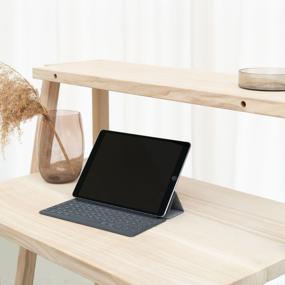 Auf einem hellen Holzregal steht ein Tablet mit schwarzer Tastatur. Daneben befindet sich eine Vase mit Trockenblumen und ein flacher, runder Glasteller. Im Hintergrund ein Vorhang in Weiß. Das Einrichtungskonzept wirkt modern und minimalistisch.