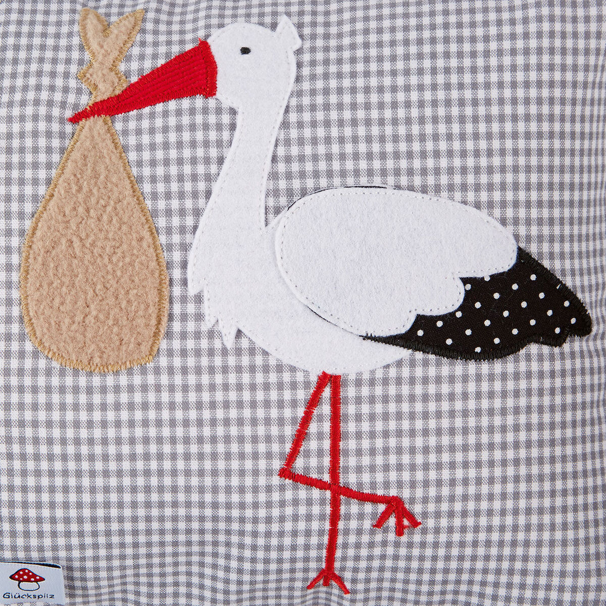 Namenskissen mit einem gestickten Storch, der ein Bündel trägt, auf grauem Karomuster-Hintergrund. Das Design umfasst die Farben Weiß, Grau, Rot, Braun und Schwarz. Geeignet als Geschenk zur Geburt, Taufe oder Babyparty.