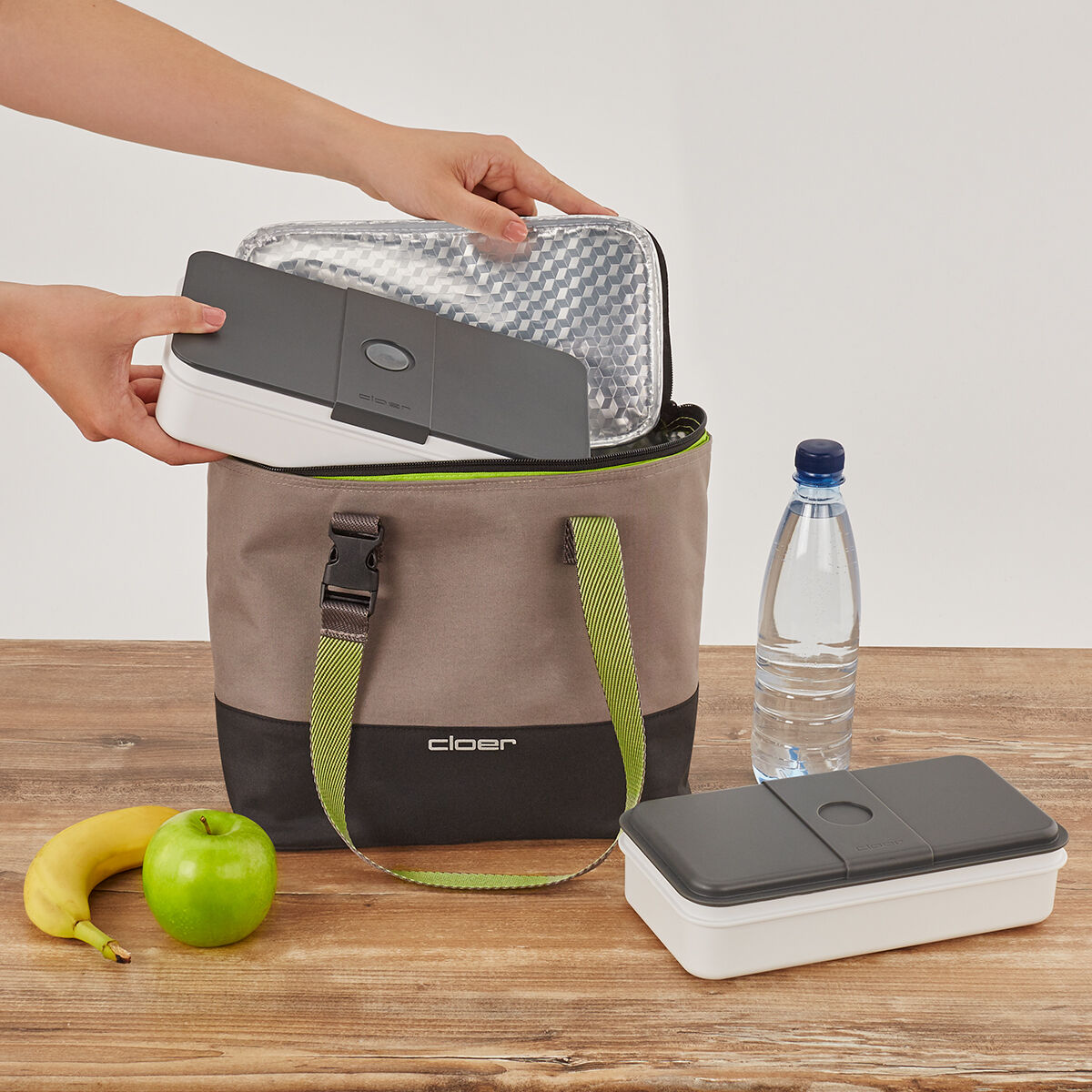 Eine Lunch-Tasche mit einer verschlossenen Lunchbox darüber. Daneben stehen eine Flasche Wasser, ein grüner Apfel und eine Banane auf einem Holztisch.