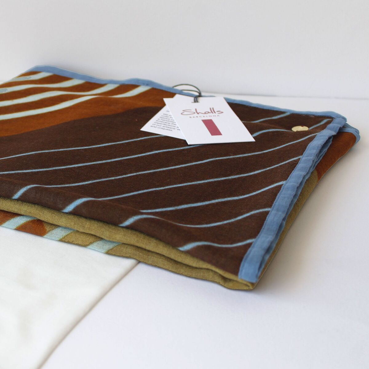 Gefalteter Wollschal 'Zelda' mit braunem und olivgrünem Streifenmuster und hellblauem Rand liegt auf einem weißen Stoffbeutel. Rechts oben befindet sich ein Etikett mit Produktinformationen. Das Bild zeigt einen modernen, modischen Schal als mögliches Geschenk oder warmes Accessoire für kalte Tage.