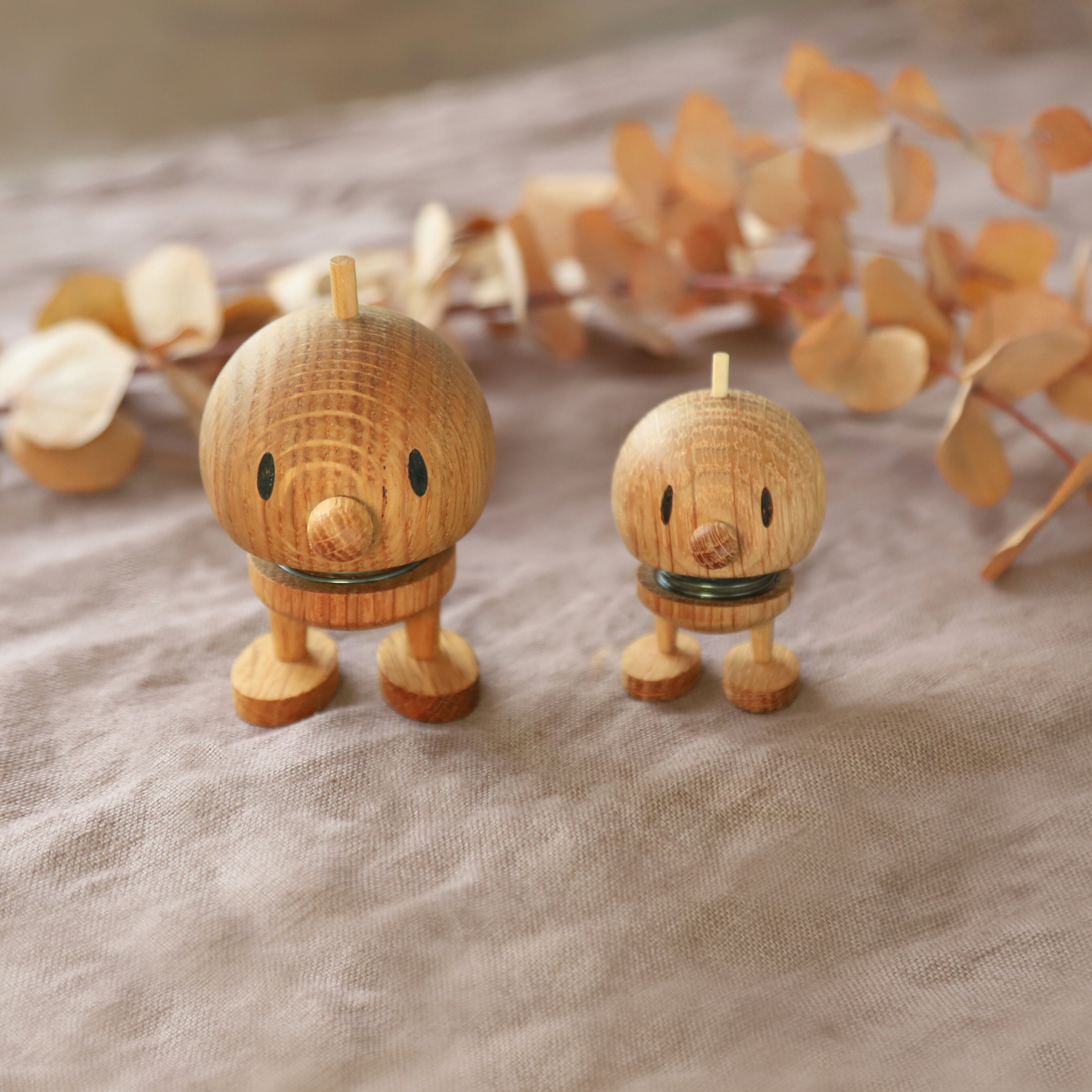 Zwei Hoptimisten der ZEIT Sonderedition aus Eichenholz, einer in Mini- und einer in S-Größe, stehen auf einem beigen Stofftuch. Im Hintergrund liegt ein Zweig mit getrockneten Blättern. Die Figuren haben freundliche Gesichter und wirken dekorativ in einer natürlichen Umgebung.