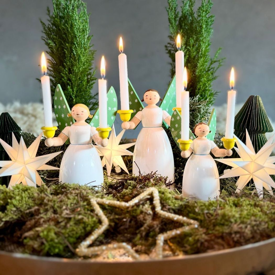 Drei traditionelle Holzfiguren ‚Lichterengel‘ von Wendt & Kühn mit weißen Kleidern, grünen Flügeln und gelben Kerzenhaltern stehen vor grünen Zweigen und leuchtenden Sternen. Alle Engel tragen weiße Kerzen, umgeben von Moos und goldenen Sternen.