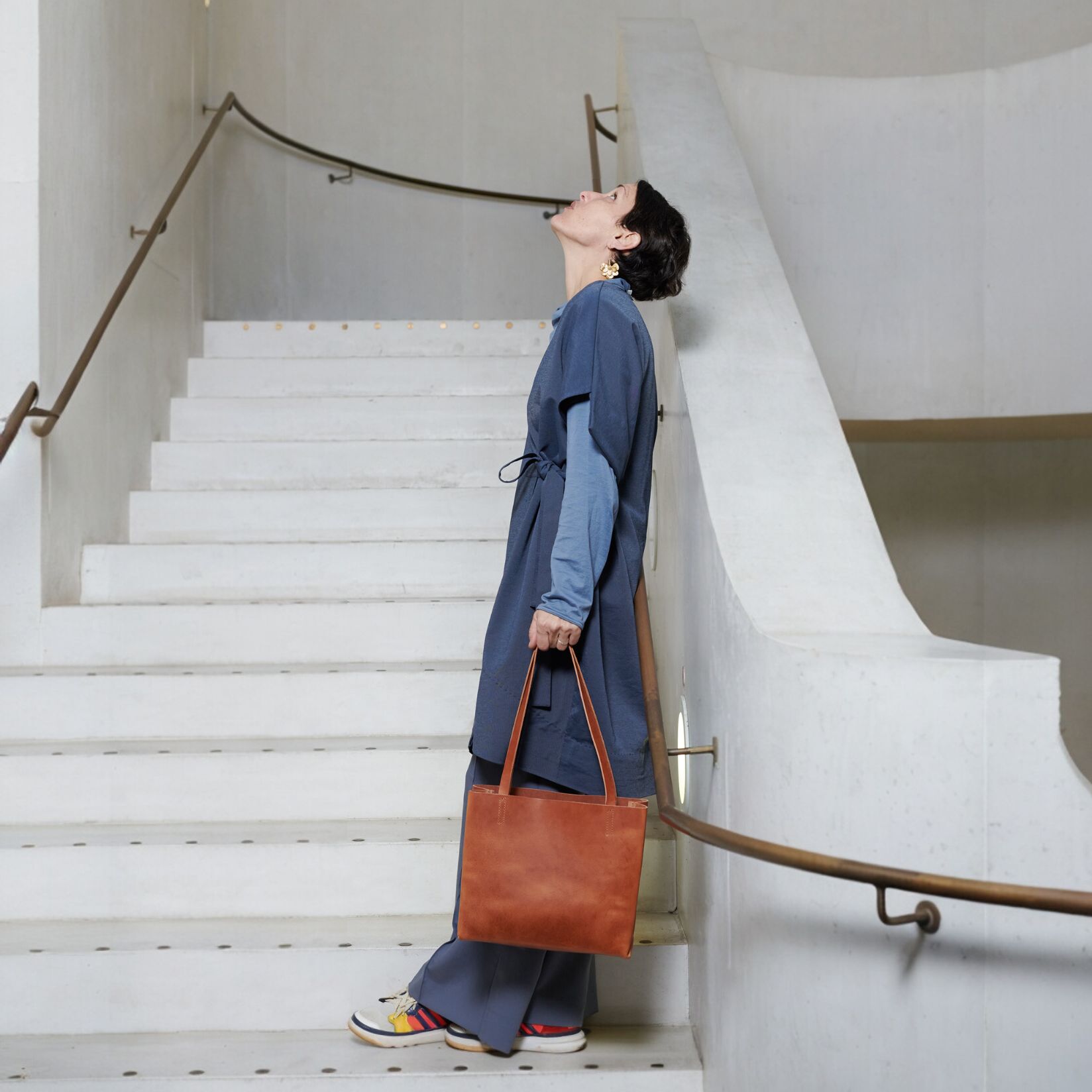 Eine Frau trägt eine braune Tasche im Paperbag-Stil an der Hand, während sie eine moderne Treppe mit geschwungenem Handlauf in einem hellen, minimalistischen Gebäude hinaufgeht. Sie trägt ein langes, locker fallendes blaues Kleid mit Sneakers. Das Bild hebt das schlichte und stilvolle Design der Tasche hervor.