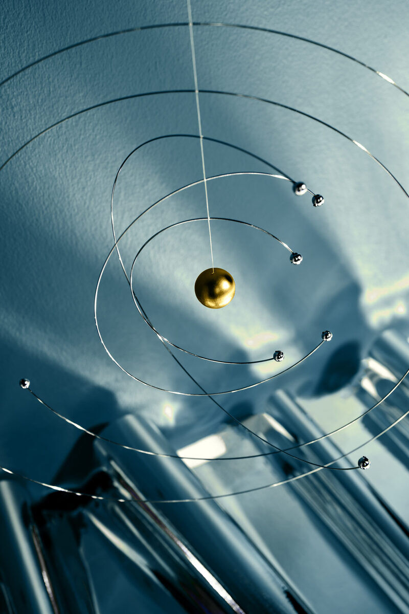 Eine hängende Skulptur aus Metallringen, die an das Niels-Bohr-Modell erinnert. Die geometrischen Formen sind zusammengefügt, um eine elegante und minimalistische Dekoration zu schaffen. Primärfarben sind Silber, Gold und Blau.