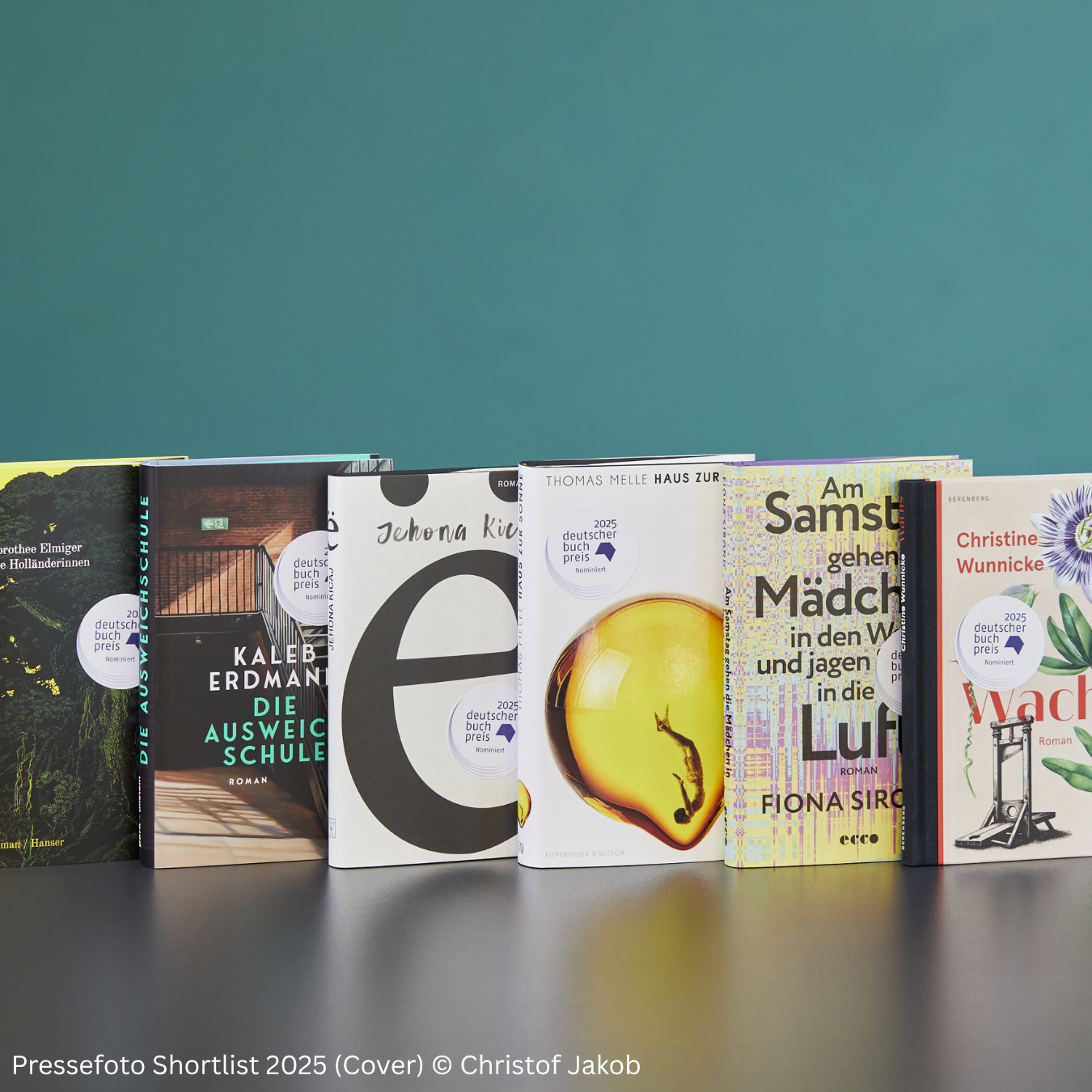 Sechs Bücher mit unterschiedlichen, farbenfrohen Covern stehen nebeneinander auf einem dunklen Tisch vor einer türkisfarbenen Wand. Auf jedem Buch befindet sich ein runder Aufkleber mit der Aufschrift „2025 deutscher buchpreis Nominiert“. Die Buchtitel sind teilweise lesbar, darunter „Die Ausweichschule“, „Haus zur Sonne“, „Am Samstag gehen wir in die Luft“ und „Wach“. Unten links steht der Hinweis „Pressefoto Shortlist 2025 (Cover) © Christof Jakob“.