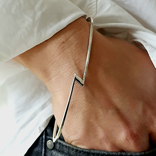 Handgelenk mit schmalem, silbernem Armreif in Zickzackform über weißem Hemdärmel und dunkler Hose.