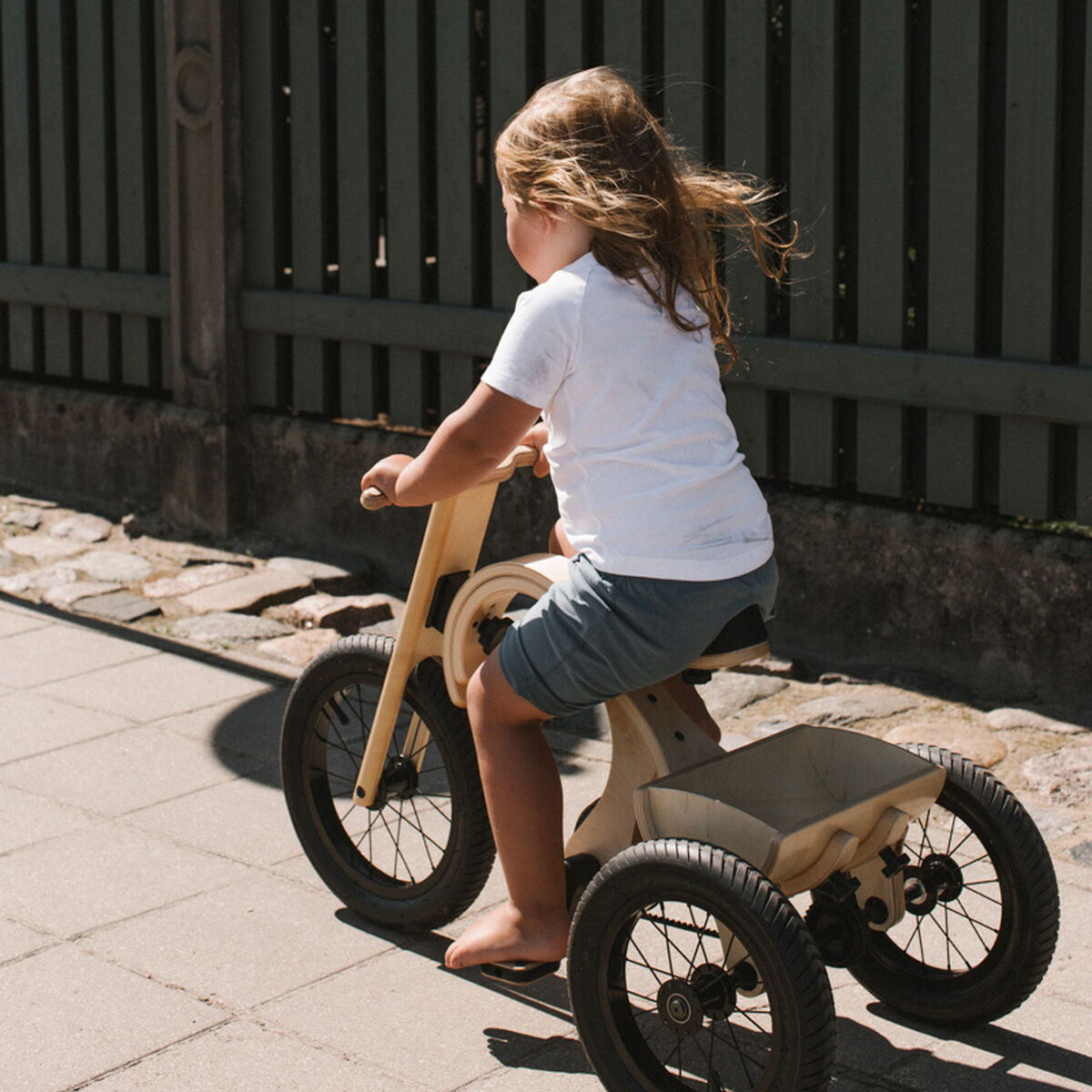 Ein kleines Kind fährt auf einem beigefarbenen Holz-Dreirad mit drei schwarzen Rädern auf einem Bürgersteig vor einem grauen Gartenzaun. Das Dreirad ist eine Erweiterung für ein Balance Bike und eignet sich für den Außengebrauch.