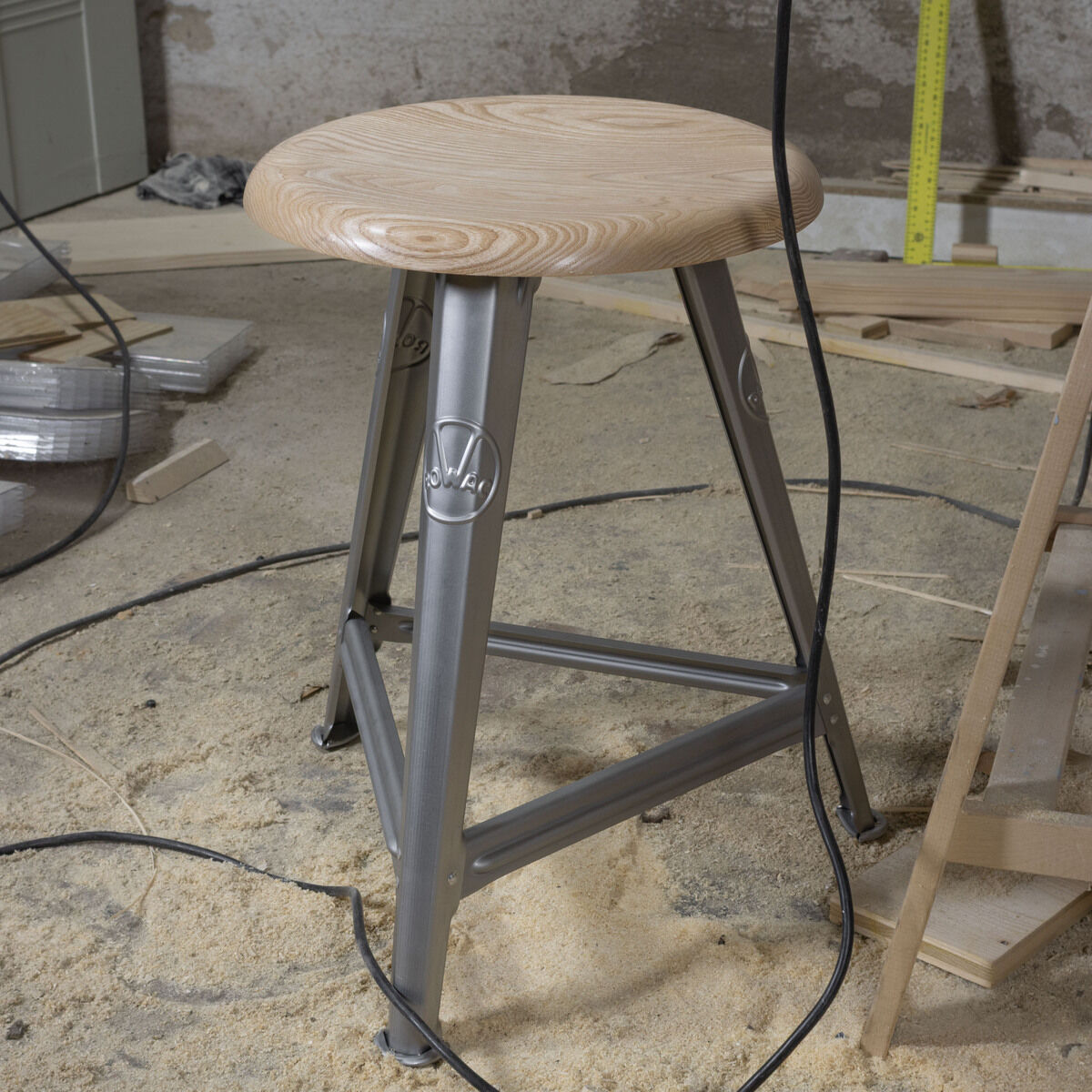 Ein stabiler Hocker mit rundem Sitz aus heller Esche und Untergestell aus blankem Stahl steht auf einem mit Sägemehl bedeckten Werkstattboden, umgeben von Werkzeugen und Baumaterialien.